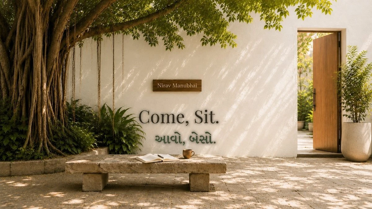 A quiet courtyard with a banyan tree, a stone bench with an open book and cup of tea, Come Sit written on a white wall in English and Gujarati, and a door standing open.