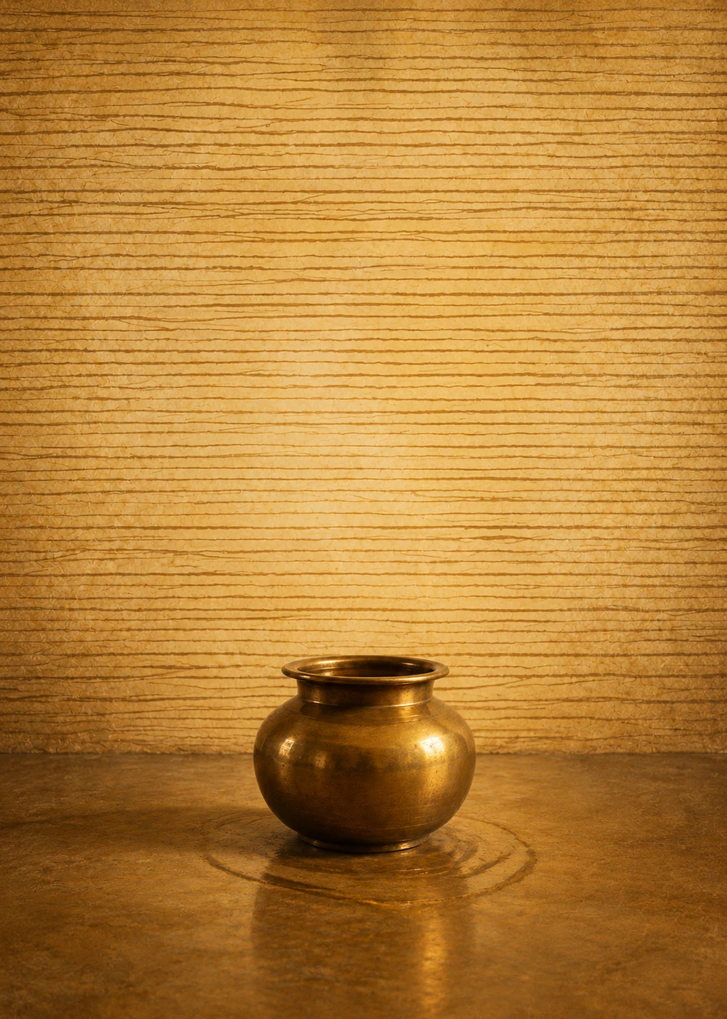 A brass vessel on a dark table, lit warmly against a pale wall.